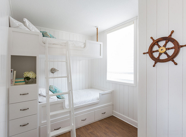 White and turquoise bunkroom. Nautical kids' bunkroom design features walls clad in vertical shiplap lined with built in bunk beds, with top bunk suspended by ropes from the ceiling, dressed in white bedding and turquoise pillows fitted with a white ladder and a built-in nook filled with books and flower stacked over a built-in dresser. #BunkRoom #WhiteBunkRoom #TurquoiseBunkRoom Laura U, Inc. 