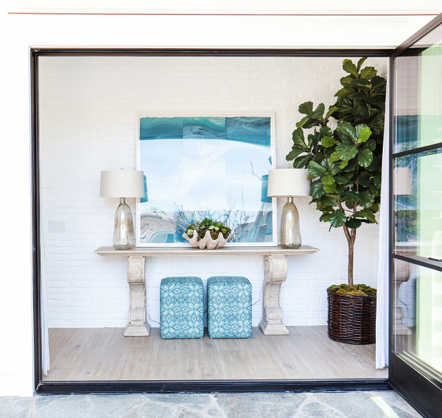 Foyer. Beach house front foyer design. Beach house coastal foyer. Folding doors open to a coastal foyer featuring a pair of turquoise stools tucked under a carved console table. Beautiful blown glass lamps and a large clam shell bowl together with a turquoise art tie this space together. Fiddle leaf fig plant. #Foyer #Turquoise #Coastal #CoastalInteriors Blackband Design.