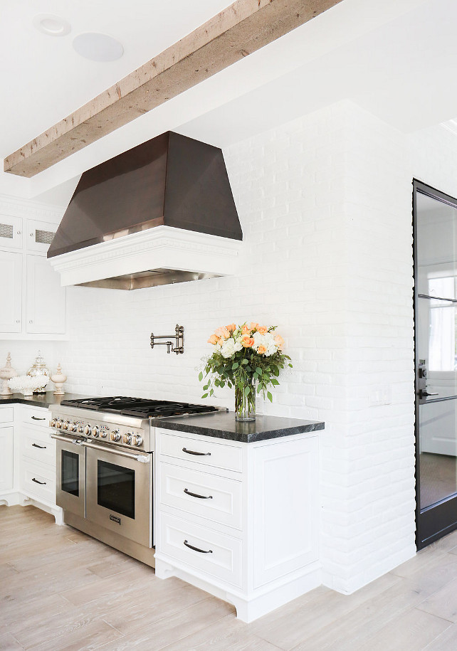 White kitchen boasts a ceiling clad in wood beams over a black and white kitchen hood which stands over a swing arm pot filler and a high end stainless steel stove. Kitchen with white cabinets paired with black marble countertops and a white brick wall backsplash alongside a gray wood floor. White Brick Kitchen Wall and Backsplash Blackband Design.