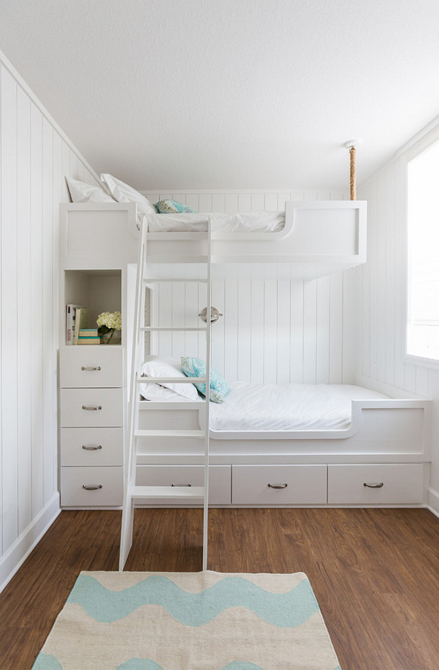 Nautical kids' bedroom design features walls clad in vertical shiplap lined with built in bunk beds, with top bunk suspended by ropes from the ceiling, dressed in white bedding and turquoise pillows fitted with a white ladder and a built-in nook filled with books and flower stacked over a built-in dresser. Laura U, Inc. 