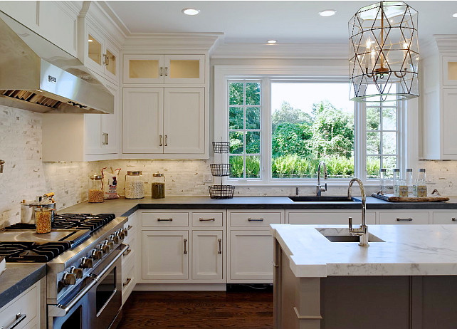 Kitchen Decor Ideas. Beautiful kitchen with white perimeter cabinets paired with honed black countertops and white marble backsplash. Worlds Away Mariah Pendant over gray kitchen island with white marble countertop with prep sink accented with gooseneck faucet. #Kitchen #KitchenDecor Blue Water Home Builders.