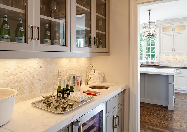 Butlers pantry. Gray butlers pantry. Butler's pantry features glass-front upper cabinets and gray lower cabinets paired with statuary marble countertop and white marble linear tile backsplash. #Butlerspantry #statuarymarblecountertop #lineartilebacksplash Blue Water Home Builders.