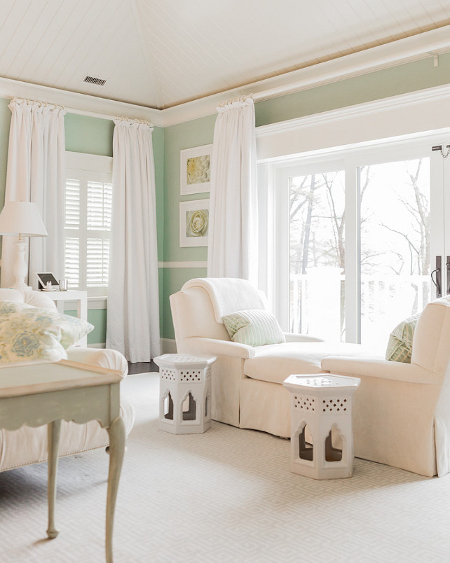 Mint Green. Mint green bedroom boasts white tongue and groove vaulted ceilings over large windows and sliding patio doors dressed in simple white drapes alongside mint green walls accented with chair rail. The bedroom features an ivory tete a tete chaise armchair topped with mint green striped pillows beside a pair of white Moroccan garden stools atop an ivory Greek key rug layered over hardwood floors. #MintGreen #Bedroom Brookes and Hill Custom Builders.