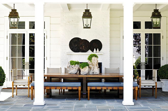 Covered Patio. Gorgeous covered patio with a pair of French doors with transom windows flanking a white brick fireplace. The outdoor patio features an iron based wood topped dining table paired with wooden benches with blue seats and upholstered side chairs. The dining table is topped with three wooden planters with green trailing plants. Three outdoor lanterns hang from the covered patios ceiling over the dining table. Traditional style planters with round boxwoods stand either side of the dining area. The outdoor dining area is finished with a flagstone patio. #CoveredPatio #Patio #Backyard Jeffrey Alan Marks.