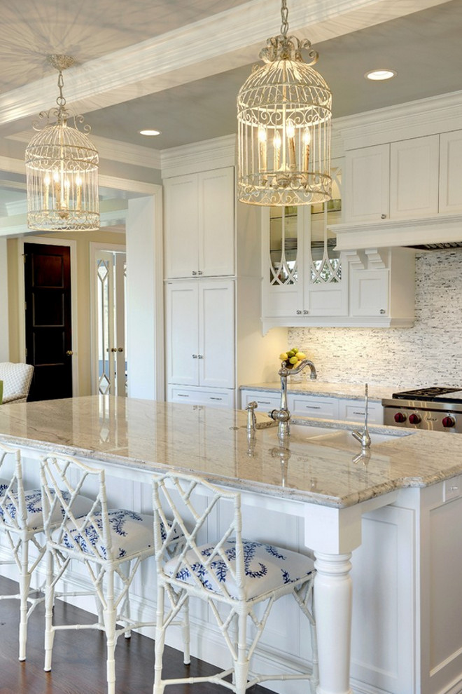 White kitchen boasts a pair of white birdcage chandeliers hanging over a white kitchen island accented with turned legs topped with gray and white granite countertops framing a white porcelain sink lined with white bamboo counter stools. Glass-front kitchen cabinets with eclipse mullion doors flank a white kitchen hood with corbels over a white and gray mosaic tiled backsplash over a high-end stainless steel stove beside full height pantry cabinets. #kitchen Stonewood Inc..jpg
