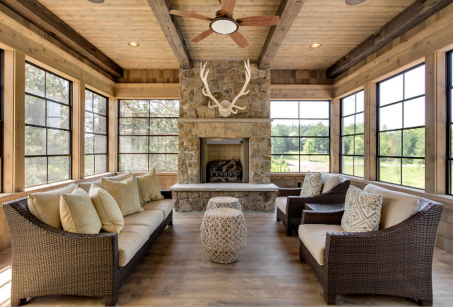 This rustic sunroom features reclaimed wood floors, reclaimed wood beams, black metal window frames and a fieldstone fireplace, Rustic Sunroom with outdoor stone fireplace antler decor Indoor Ceiling Fan recessed lighting wicker furniture wood ceiling wood walls