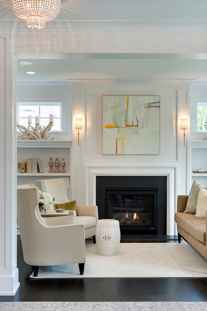 This living room features a gray and orange abstract art piece illuminated by Dauphine Sconces hanging over a white, sleek fireplace adorned with a black slate surround flanked by open built in bookcases under windows. White living room painted in Benjamin Moore OC-117 Simply white featuring neutral furniture and decor. #Livingroom #neutrallivingroom #whitelivingroompaintcolor #BenjaminMooreOC117Simplywhite #neutrallivingroompaintcolor #neutrallivingroomfurniture Spacecrafting Photography. City Homes Design and Build, LLC