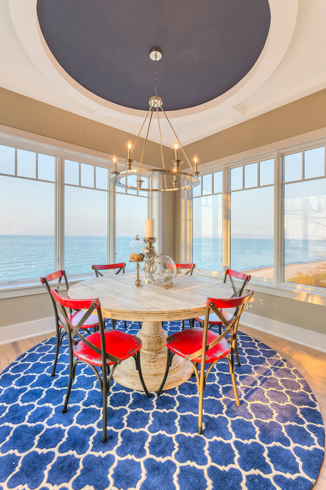 Coastal Dining Room beach front Beachfront Interiors candlesticks classic design distressed red dining chairs lakefront home large windows Nautical Luxuries navy blue ocean view round blue area rug round ceiling design round dining table sunroom water front water view