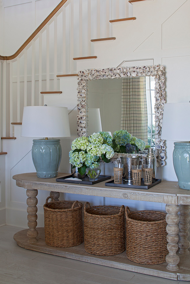 Coastal Foyer with natural elements. This coastal foyer features wainscoted staircase, an oval console table filled with three round woven baskets and topped with watery blue table lamps and a large rectangular seashell encrusted mirror. #CoastalFoyer #Foyer #Foyeswainscotting #Foyerwalls #Foyerpanelledwalls #coastalinteriors Urban Grace Interiors