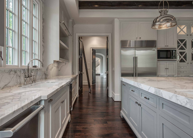 Kitchen island with gray and white marble countertop framing a corner prep sink situated across from gray open shelving flanking window over a white porcelain sink and deck-mount gooseneck faucet flanked by stainless steel dishwashers. An under cabinet stainless steel refrigerator stands beside an integrated microwave and coffee station cabinet. #kitchen #sink #graykitchen #farmhousekitchen #farmhousesink #islandsink Elizabeth Garrett Interiors.