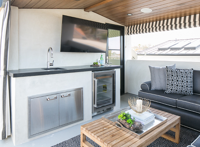 The roof deck features stained paneled vaulted ceiling over a kitchenette boasting a flatscreen tv over a sink and stainless steel cabinets next to a glass-front beverage fridge. Patterson Custom Homes