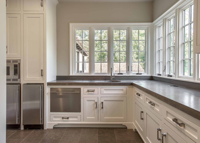 Butler's pantry. Large butlers pantry features creamy white shaker cabinets fitted with a pull-out dishwasher drawer topped with gray countertops situated under windows next to a stainless steel microwave stacked over stainless steel mini fridges. #butlerspantry #largebutlerspantry Elizabeth Garrett Interiors.