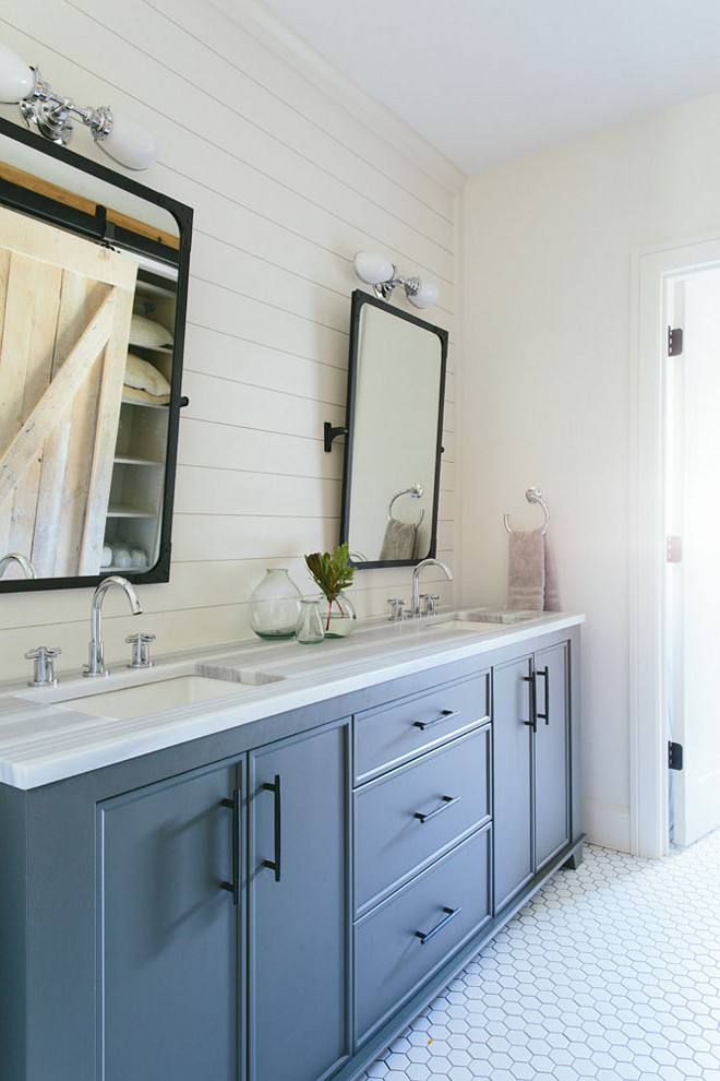 Blue gray bathroom cabinet. Bathroom features blue gray bathroom cabinets and a shiplap accent wall lined with a blue gray double washstand adorned with long bronze pulls topped with striped marble fitted with two sinks and polished nickel high arc faucets placed under Restoration Hardware Industrial Rivet Pivot Mirrors lit by Vintage English Oval Double Sconces alongside a white hex tiled floor finished with white grout. Kate Marker Interiors.