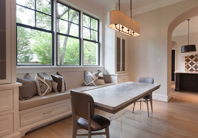 Breakfast nook. Breakfast nook boasts a long built in window seat bench lined with gray and black pillows flanked by metal lattice china cabinets. A window seat dining bench faces an acrylic based dining table with stone top lined with gray herringbone chairs illuminated by an oval linen pendant light. Elizabeth Garrett Interiors