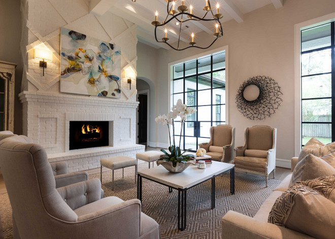 White and beige living room features a high ceiling dotted with white wood beams and a black and gold curvy chandelier illuminating a pair of burlap french wingback chairs facing a pair of linen tufted chairs facing each other across from a stone top coffee table atop a beige diamond woven rug. A pair of white linen stools sit in front of a white brick fireplace and hearth lined with a blue and gold abstract canvas art piece illuminated by iron wall sconces. Elizabeth Garrett Interiors