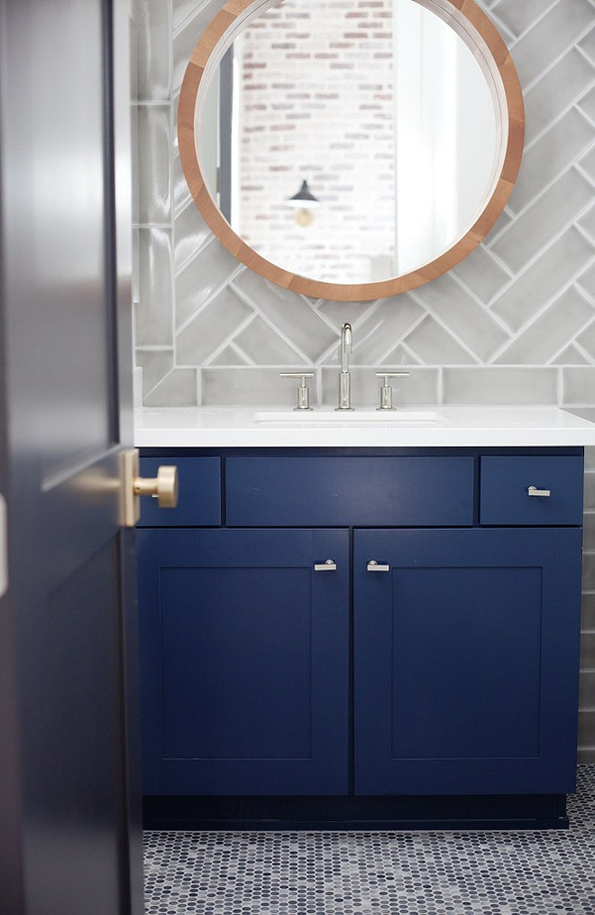 Bathroom. Transitional Blue and gray bathroom boasts a round mirror lining a glossy gray chevron tile backsplash placed above a bold blue cabinet with white quartz countertop. #Bathroom #Transitional #interiors #bluecabinet #whitequartz E Interiors
