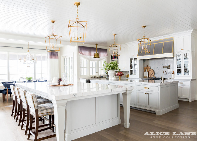 Ivory Lane Kitchen. A cook's kitchen features a white beadboard ceiling dotted with four Darlana 4 Light Lanterns illuminating two kitchen islands topped with white marble with one island designated for prep work and the other island designated for a dining table lined with gray check counter stools. A steel kitchen hood is accented with brass trim and stands over a marble cooktop backsplash lined with a swing arm pot filler and a stainless steel stove with dual ovens. A farmhouse sink fitted with aged gooseneck faucets is flanked by stainless steel dishwashers placed under windows dressed in a purple ombre roman shade illuminated by a Single Sloane Shop Light with Metal Shade. #kitchen #ivorykitchen #twoislands #IvoryLaneKitchen Alice Lane Home.