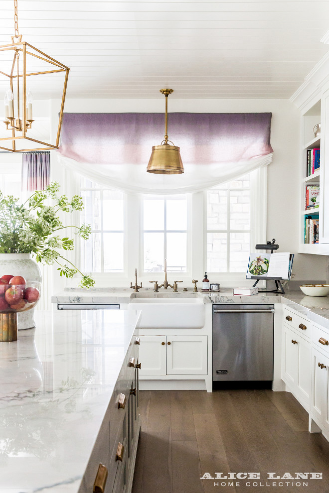 Kitchen Lighting. Brass Kitchen Lighting. Farmhouse Sink with Aged Brass Vintage Faucets Placed Under Antique Brass Drum Pendant Light. kitchen features a farmhouse sink fitted with aged gooseneck faucets is flanked by stainless steel dishwashers placed under windows dressed in a purple ombre roman shade illuminated by a Single Sloane Shop Light with Metal Shade. #kitchen #lighting #farmhousesink #kitchenlighting Alice Lane Home