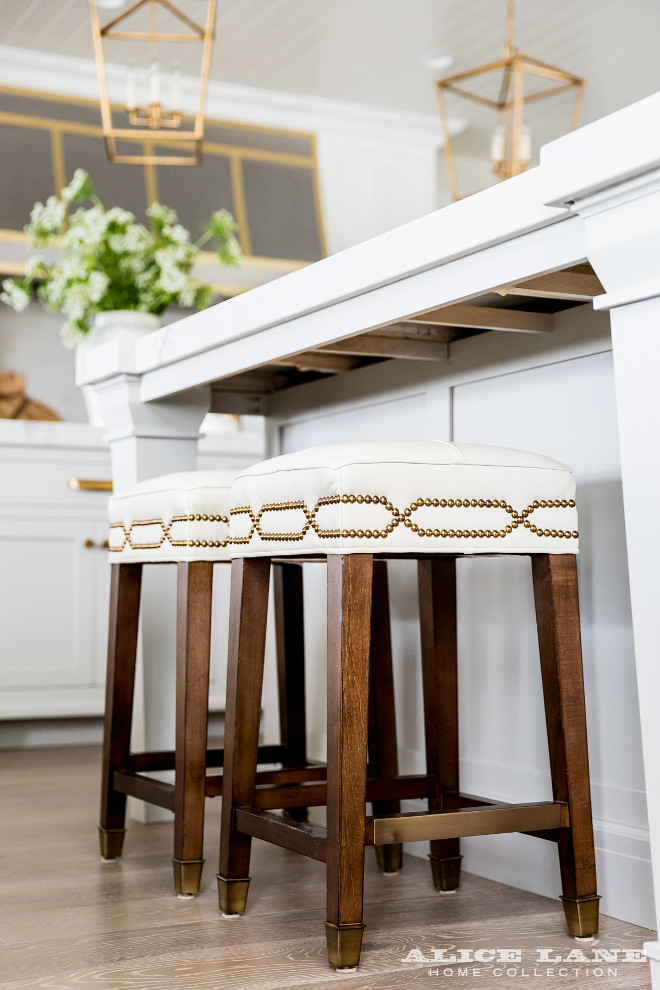 Kitchen stools. Kitchen features a white center island with legs lined with backless white counter stools with brass nailhead trim illuminated by Darlana Medium Lanterns in Antique Brass. #KitchenStools #stools #counterstools #barstools Alice Lane Home.