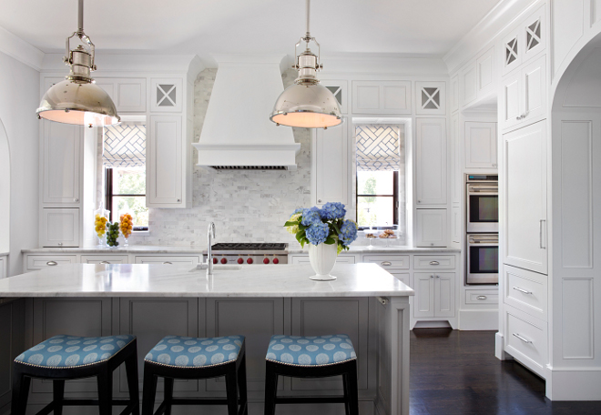 White kitchen with gray island. This kitchen features light gray kitchen island with marble countertop and small prep sink lined with backless black bar stools upholstered in blue medallion fabric. The kitchen also features crisp white shaker cabinets paired with marble subway tile backsplash. Steel windows covered in white and gray geometric roman shades flank white paneled kitchen hood over a Wolf Range. #whitekitchen #white #kitchen #grayisland #kitchenisland TS Adams Studio Architects. Traci Rhoads Interiors.