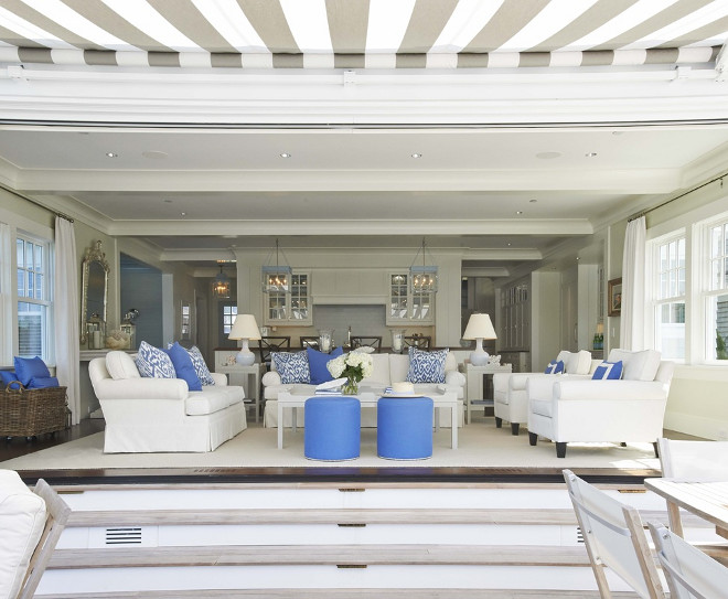 Living Room. White and blue living room features white roll-arm chairs adorned with blue border pillows facing white roll-arm loveseat across from white coffee table and round blue stools with silver nailhead trim atop white rug layered over espresso wood floors. A white roll-arm sofa is adorned with white and blue ikat pillows is flanked by gray end tables topped with ceramic lamps facing open doorway and steps leading to patio. Lynn Morgan Design.