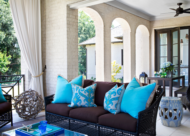Porch. Back porch. The back porch features off-white brick walls and columns accented with white outdoor curtains. Notice the chocolate brown rattan sofa with brown cushions, turquoise ikat pillows and turquoise blue pillows. #porch #backporch TS Adams Studio Architects. Traci Rhoads Interiors. 