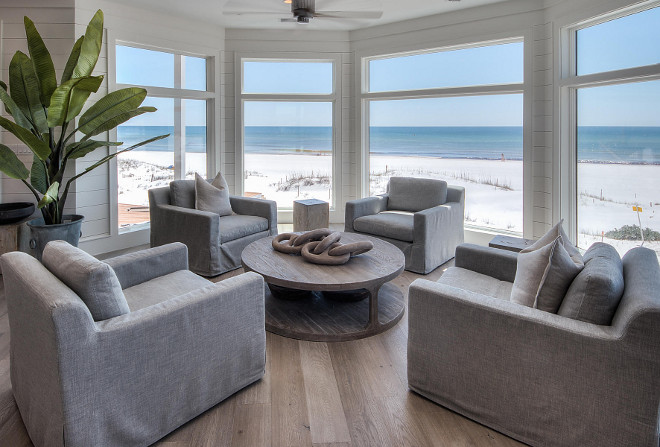 Transitional Coastal Interiors. Just off the living room, a sitting area is nestled in front of a gallery of bay windows. Beach house with Transitional Coastal Interiors. Beach house with transitional coastal interiors, with whitewashed plank hardwood floors, slipcovered furniture, reclaimed furniture and tongue and groove walls. #TransitionaInteriors #Interiors #CoastalInteriors