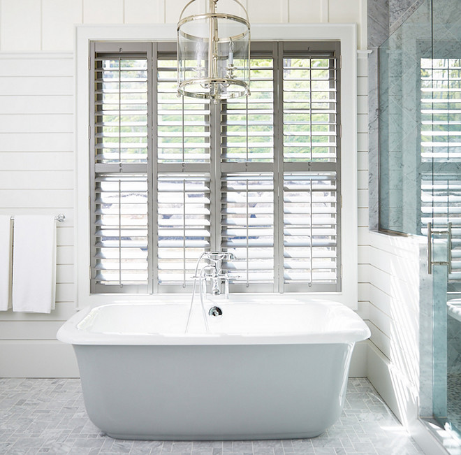 Bathroom Window Shutter Ideas. Bathroom features a Round Edwardian Entry lantern suspended over a freestanding tub placed under windows dressed in plantation shutters alongside a grey marble tiled floor next to a shiplap and glass walk in shower. Muskoka Living