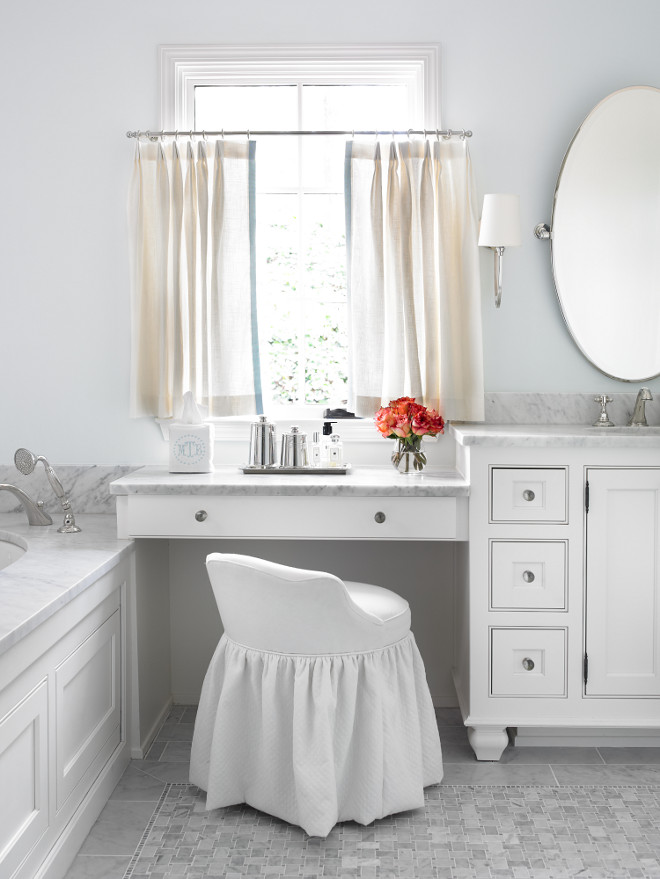 Make up vanity in bathroom. Bathroom with marble-top drop down make-up vanity under window covered in linen cafe curtains paired with white slipcovered vanity stool. White bathroom cabinets with carrara marble countertops and oval pivot mirror accented with monogrammed embroidered tissue box cover. Master bathroom features wood paneled drop-in tub with carrara marble surround over carrara marble mosaic tile floor. L. Kae Interiors