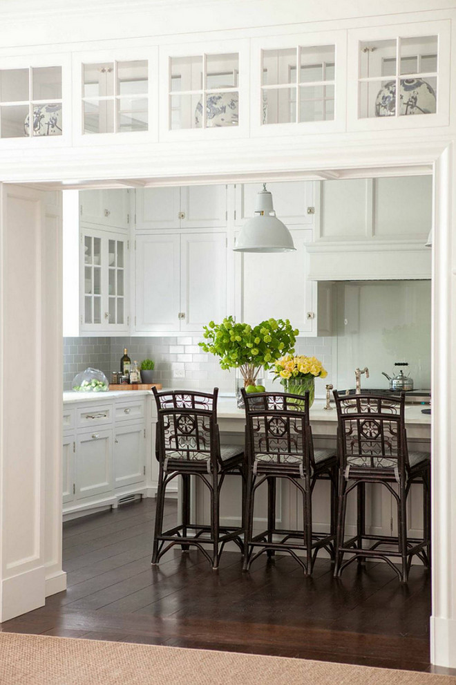 Kitchen Doorway. A kitchen doorway is fitted with glass display cabinets leading to a beautiful kitchen filled with white shaker cabinets paired with white marble countertops and a gray subway tile backsplash. A pair of white industrial pendants hang over a white center island fitted with a sink lined with brown bamboo counter stools by Redegg Indochine Bar Stools. Redegg Indochine Bar Stools. Dibico Construction