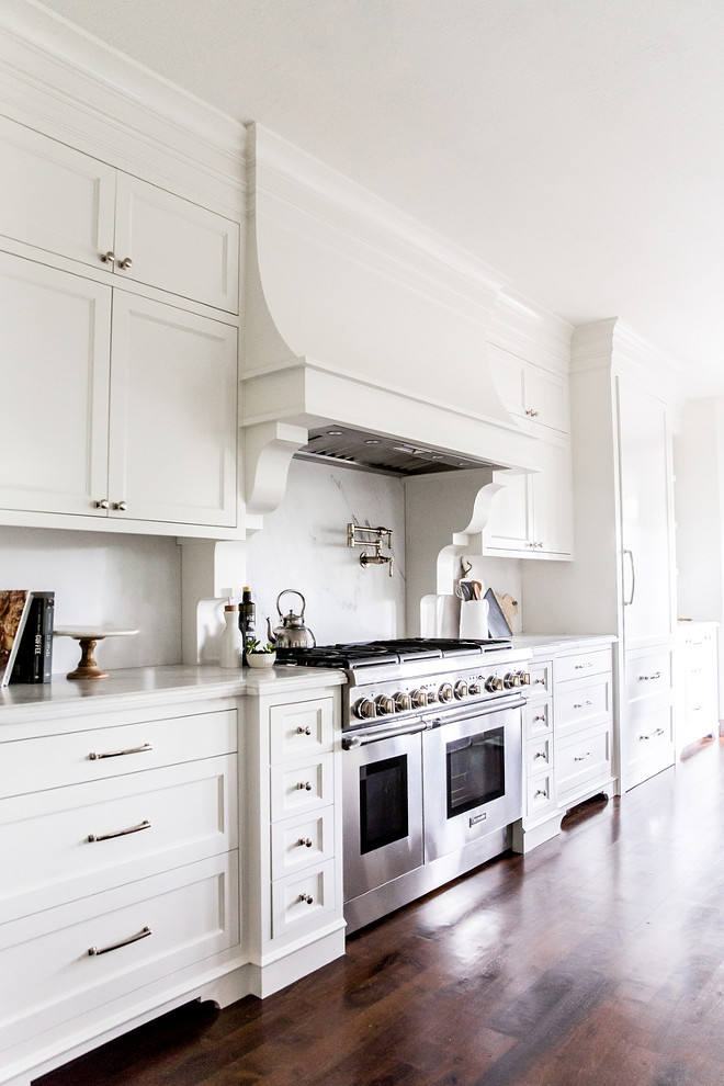White French Kitchen Hood with Corbels and White Marble Slab Backsplash. White kitchen features white shaker cabinets paired with white marble countertops with gray veining and matching backsplash. A white French kitchen hood with corbels stands over a swing arm pot filler and a stainless steel stove.. white-french-kitchen-hood-with-corbels-and-white-marble-slab-backsplash White French Kitchen Hood with Corbels and White Marble Slab Backsplash #WhiteFrenchKitchen #FrenchHood #Corbels #WhiteMarbleSlab #Backsplash LIV Design Collective.