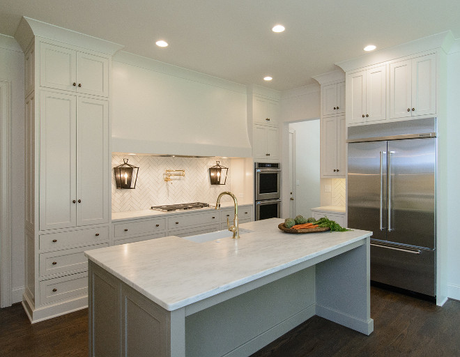 Clean lined kitchen. Cabinet Perimeter Paint Color: Shoji White by Sherwin Williams. Island: Sherwin Williams Intellectual Gray. This clean-lined white kitchen boasts a stainless steel refrigerator recessed beneath white cabinets facing a gray island topped with a white quartz countertop fitted with a farmhouse sink with a brass gooseneck faucet. A extra long white hood is mounted above a brass pot filler flanked by iron wall lanterns. The white herringbone backsplash tile creates some texture in this space. Clean lined kitchen ideas. Clean lined kitchen #Cleanlinedkitchen #PerimeterPaintColor #ShojiWhiteSherwinWilliams #SherwinWilliamsIntellectualGray Tasha B. Davis Interiors, LLC.