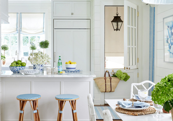  White Kitchen Peninsula with Backless Blue Bistro Stools. Beautifully designed white kitchen with light blue accents boasts Serena & Lily Riviera Backless Stools fitted with light blue cushions placed in front of a white peninsula topped with a white marble countertop while a blue and white is painting mounted on a shiplap wall beside a white glass paneled Dutch door positioned next to an under cabinet white paneled fridge. The fridge is located beside windows covered in white and blue roman shades positioned over an apron sink paired with a polished nickel deck mount gooseneck faucet. White Kitchen Peninsula with Backless Blue Bistro Stools. cottage-kitchen Sarah Bartholomew Design