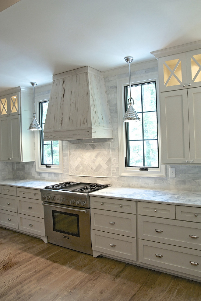 Grey kitchen. Stunning transitional kitchen is equipped with a rustic pecky cypress hood fixed between windows framed by gray marble backsplash tiles illuminated by industrial pendants. The hood is located above herringbone marble cooktop tiles accenting a Thermador range flanked by white drawers with polished nickel cup pulls topped with gray and white marble countertops positioned beneath white upper cabinets finished with x front lighted display cabinets. According to the designer, the cabinet paint color was a custom color, but the closest color would be White Dove by Benjamin Moore. grey-kitchen Tasha B. Davis Interiors, LLC.