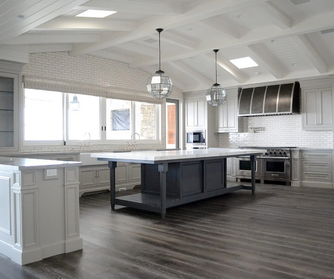 Kitchen. This large kitchen features a freestanding blue grey island with white marble countertop, illuminated by Visual Comfort Morris pendant lights hung from a vaulted shiplap ceiling fitted with multiple skylights. The island sits in front of a wall of windows framed by white linear tiles fixed above light gray cabinets. To the side, a black barrel hood is mounted on white linear tile backsplash above a Wolf dual range. Flooring is a stained oak floor. #kitchen #largekitchen #freestandingisland #whitemarblecountertop #VisualComfortMorris #pendantlights #vaultedshiplapceiling #wallofwindows #whitelineartiles #lightgraycabinets #blackbarrelhood Matt Morris Development.