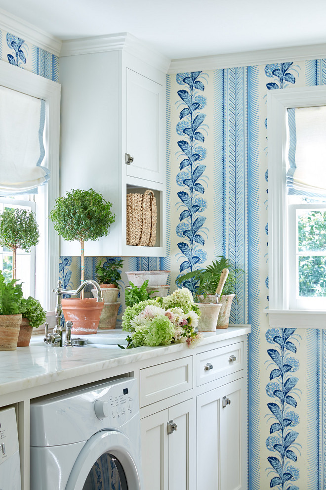  White and Blue Cottage Laundry Room with Topiaries. White and blue cottage laundry room boasts walls clad in white and blue wallpaper framing a window dressed in a blue and white roman shade as an adjacent wall is fitted with white cabinets adorning nickel hardware and finished with a white front loading washer and dryer positioned under a honed white marble countertop. A sink with a polished nickel hook and spout faucet is fixed beneath a window located beside a white upper cabinet completed with an open shelf. White and Blue Cottage Laundry Room with Topiaries #WhiteandBlue #LaundryRoom #Topiaries laundry-room Sarah Bartholomew Design