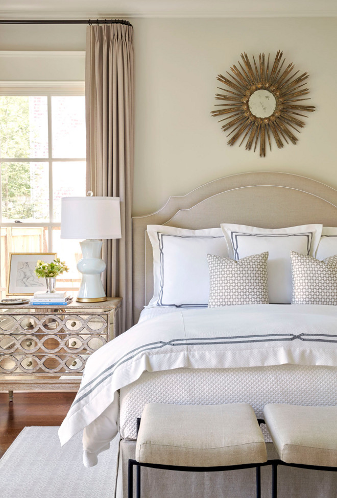 Gray Bedroom with Mirrored Nightstand Under Window. Gray bedroom features a gold sunburst mirror placed over a gray velvet headboard on bed dressed in white and gray hotel bedding as well as a linen bench placed at the end of the bed. A gray curved headboard is placed next to a mirrored nightstand, Ogee French Country Distressed Antique Mirror Dresser Chest, topped with a white lamp situated in front of a window dressed in gray French pleat drapery panels. Gray Bedroom with Mirrored Nightstand Under Window #GrayBedroom #MirroredNightstand neutral-bedroom Sarah Bartholomew Design