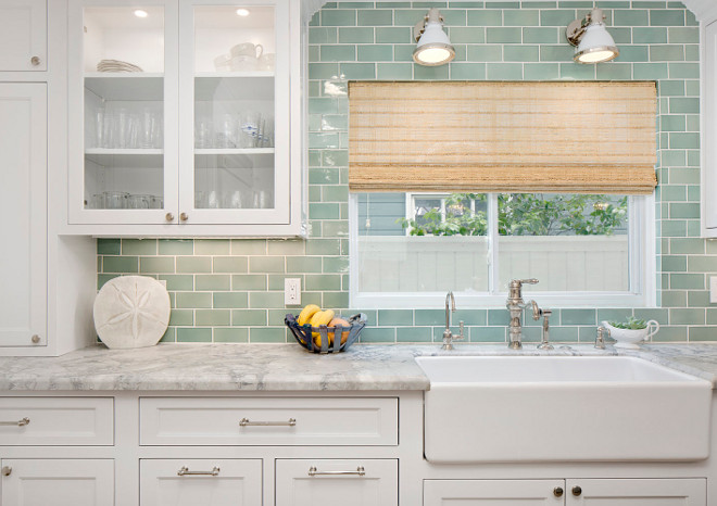 Seafoam Green Subway Tile Backsplash. Kitchen with white cabinets and Seafoam Green Subway Tile Backsplash. White industrial sconces are mounted on green subway tiles framing a window dressed in a bamboo roman shade hung above a farmhouse sink with a polished nickel faucet fixed in a marble countertop complementing white inset cabinets finished with polished nickel pulls as upper glass front cabinets are lit by custom lighting. Seafoam Green Subway Tile Backsplash #SeafoamGreen #GreenSubwayTile #Backsplash seafoam-green-subway-tile-backsplash Erin Hedrick