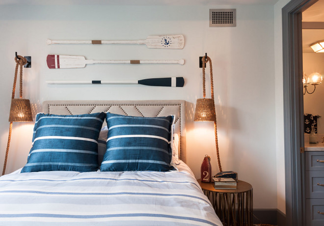 Vintage Oars Over Beige Nailhead Headboard. Cottage bedroom features vintage decorative wall oars fixed between woven swing arm rope sconces over a beige nailhead headboard accenting a bed dressed in a blue and white striped duvet topped with blue striped pillows flanked by gold nightstands. #VintageOars #NailheadHeadboard Matt Morris Development