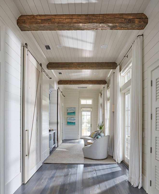 Floor to ceiling shiplap paneling with reclained wood beam. This hallway boasts rustic wood beam ceiling, shiplap walls, shiplap ceiling as well a wall of French doors and transom windows dressed in white cotton curtains. A chic hall boasts a nook filled with a blue wet bar flanked by rooms finished with pecky cypress barn doors on rails. #shiplap #reclaimedwood #floortoceilingshiplap floor-to-ceiling-shiplap-paneling-with-reclaimed-wood-beam