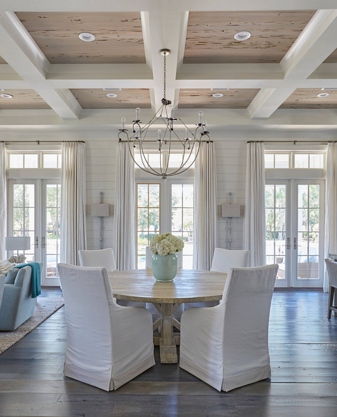 Dining Room Coffered Ceiling with Pecky Cypress Trim. The dining room boasts a glossy white coffered ceiling accented with pecky cypress coffers accented with an iron chandelier. The round salvaged wood dining table surrounded by white slipcovered dining chairs. Lighting is Lowcountry Originals Spring Island Basket. #lowcountryoriginals #lighting #springislandbasket #chandelier #DiningRoom #CofferedCeiling #PeckyCypress #Trim