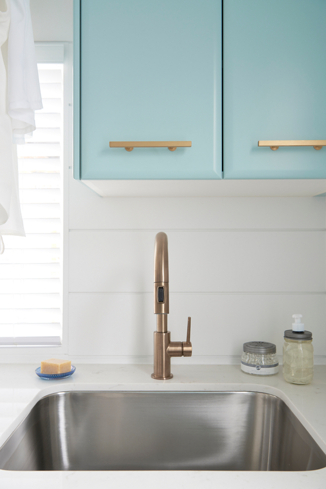 Brass Faucet. Laundry room brass faucet. Faucet is Delta Trinsic Single Handle Pull-Down Kitchen Faucet in Champagne Bronze. A curved stainless steel sink paired with a gold gooseneck faucet is fixed to a white quartz countertop beneath blue laundry room cabinets, painted in Gossamer Blue by Benjamin Moore, accented with brass pulls, and mounted beside a window. #laundryroomfaucet #faucet Soda Pop Design Inc.