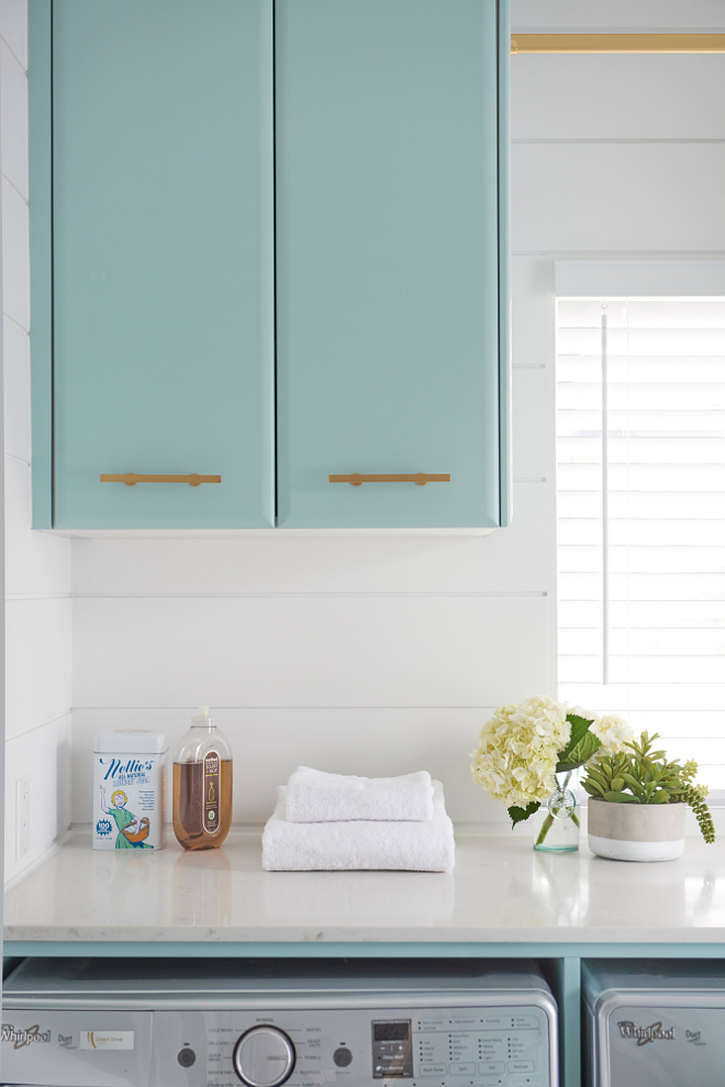 Laundry room quartz laundry room. Countertop: Simplex Marble & Granite - Cashmere Carrara. Blue laundry room cabinets painted in Gossamer Blue by Benjamin Moore are accented with brass pulls and mounted beside a window against a white shiplap trim lining a white quartz countertop positioned above an enclosed silver washer and dryer #Laundryroom #quartz #laundryroom #countertop Soda Pop Design Inc.