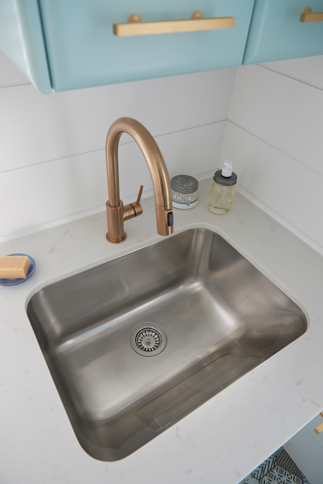 Well appointed contemporary laundry room boasts a curved stainless steel sink fixed to a white quartz countertop and paired with a gold gooseneck faucet mounted in front of a white shiplap trim beneath blue cabinets painted in Gossamer Blue by Benjamin Moore and finished with brass pulls. #laundryroom Soda Pop Design Inc.