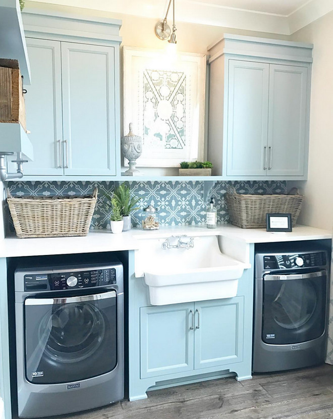 Laundry room with sink. Laundry room sink. Laundry room features Blue gray cabinets and a farmhouse sink flanked by a pair of gray washer and dryer. Countertop is white quartz and backsplash is patterned cement tile. #laundryroom #bluegraycabinets #laundryrooms #cementtile #patternedtile #quartzcountertop #sink #farmhousesink #laundryroomsink Artisan Signature Homes
