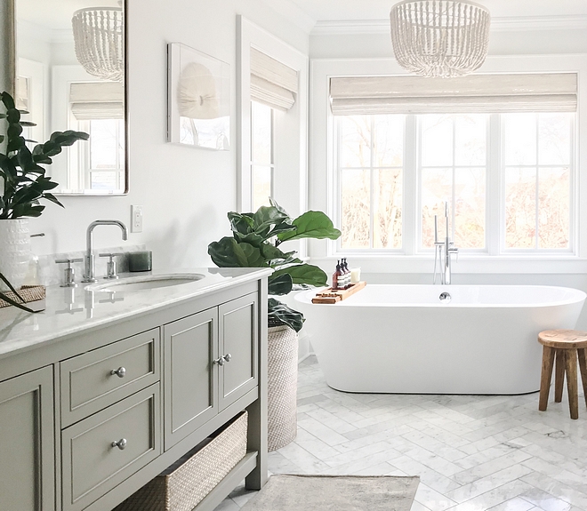 Bathroom featuring herringbone marble floor tile, white beaded chandelier over freestanding tub surrounded by windows dressed in white bamboo Roman shades #bathroom #herringbonemarbletile #whitebeadedchandelier #beadedchandelier #bambooromanshade #romanshade #freestandingtub #masterbathroom