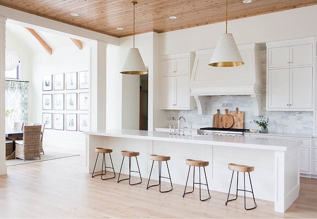 A pair of white and gold cone pendants, Goodman Hanging Lamps, illuminates an extra-long center island topped with white quartz fitted with a farmhouse sink and deck mount faucet lined with wood and iron counter stools, Wisteria Smart & Sleek Stools. A white kitchen hood with corbels stands over a marble brick tiled backsplash and a stainless steel stove. KitchenKitchen Ashley Winn Design.