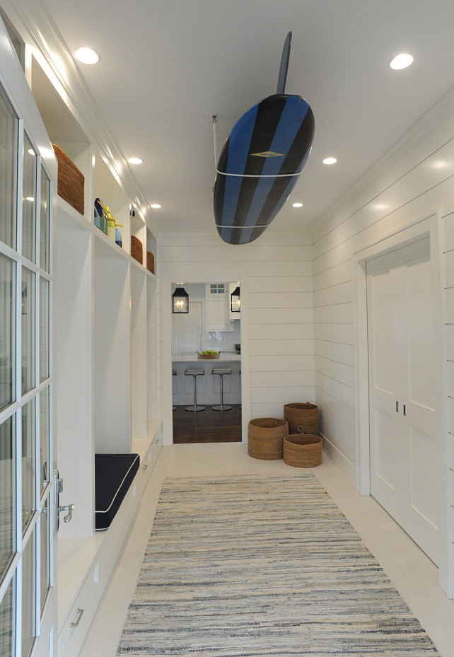 Mudroom. Coastal Mudroom. Coastal Mud Room Ideas. Beach mud room with wall length, built-in lockers featuring a built-in storage bench with overhead cubbies alongside wood planked walls with a ceiling mount surfboard over ivory floors layered with a gray rag rug. #Mudroom #Coastal Nina Liddle Design.