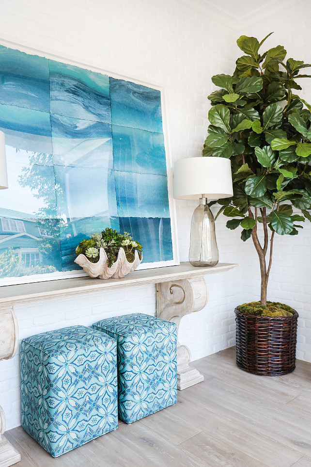 Turquoise Foyer. Coastal foyer with turquoise decor. Beautiful foyer features a pair of turquoise stools tucked under a carved console table topped with smoke gray blown glass lamps and a large clam shell bowl under turquoise art alongside a fiddle leaf fig plant. #Foyer #Turquoise #CoastalInteriors Blackband Design.
