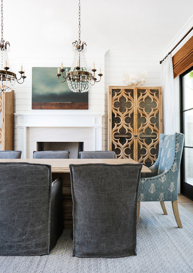 Dining Room Design. This gorgeous dining room features a pair of Paris flea market chandeliers over a wood dining table lined with charcoal gray slipcovered dining chairs as well as blue wingback dining chairs. You will also notice the shiplap accent wall and fireplace flanked by quatrefoil glass cabinets. The glass and steel doors are dressed in bamboo roman shades and white linen draperies. #DiningRoom